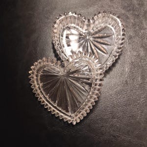 May include: Two clear, heart-shaped glass dishes with a starburst design in the center. The edges are scalloped. The dishes are on a dark surface. These decorative dishes are ideal for small trinkets.