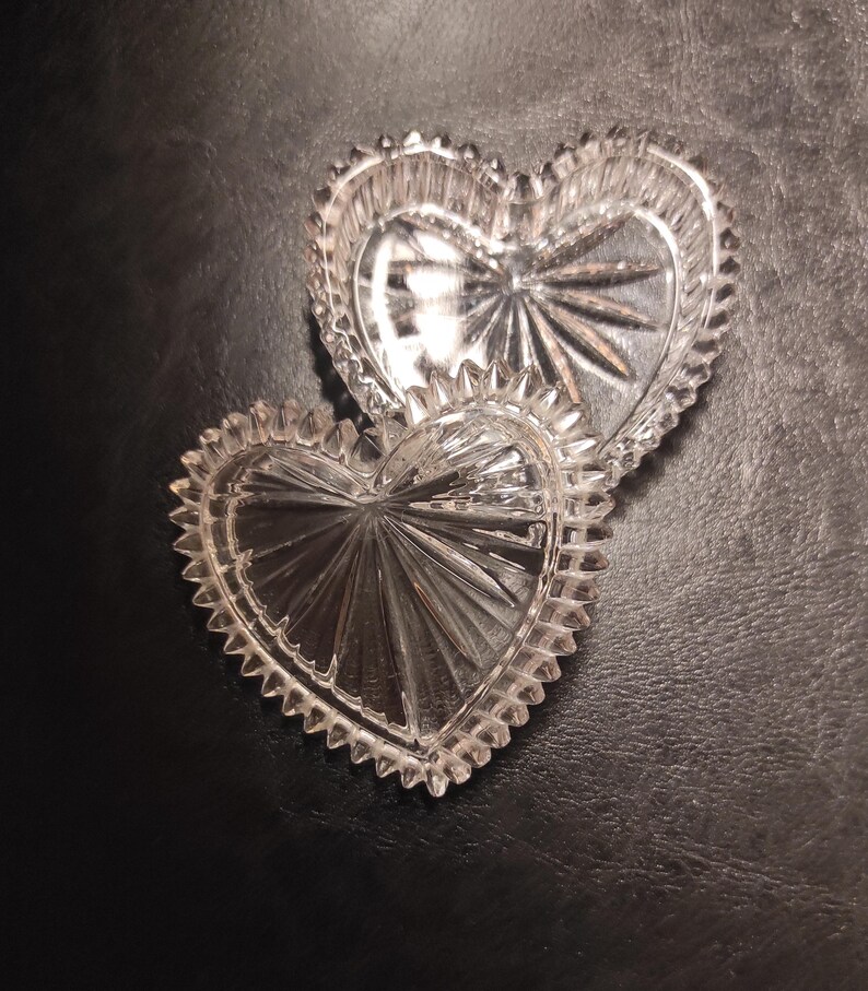 May include: Two clear, heart-shaped glass dishes with a starburst design in the center. The edges are scalloped. The dishes are on a dark surface. These decorative dishes are ideal for small trinkets.