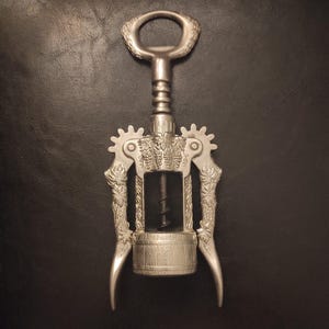 May include: Silver wine corkscrew with decorative detailing. The corkscrew has a handle, a worm, and two levers. The design incorporates grape clusters and leaf patterns. The metal corkscrew is designed for opening wine bottles.