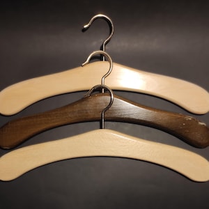 Puede incluir: Tres perchas de madera con ganchos de metal. Las perchas superior e inferior son de color beige claro, mientras que la percha del medio es de color marrón oscuro. Las perchas están diseñadas con una forma de hombro ancha.