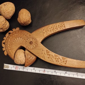 Op de afbeelding: Een houten notenkraker met ingewikkelde houtsnijwerk. De notenkraker is gevormd als een vogel met een groot, rond hoofd en een lange, dunne snavel. De notenkraker is open, waardoor een walnoot zichtbaar is. Een liniaal ligt onder de notenkraker.