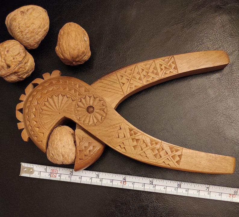 Op de afbeelding: Een houten notenkraker met ingewikkelde houtsnijwerk. De notenkraker is gevormd als een vogel met een groot, rond hoofd en een lange, dunne snavel. De notenkraker is open, waardoor een walnoot zichtbaar is. Een liniaal ligt onder de notenkraker.