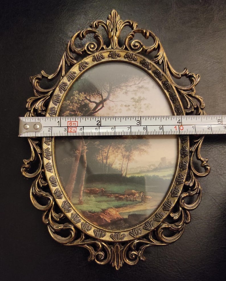 Puede incluir: Marco de fotos ovalado y ornamentado con una pintura de paisaje. El marco es de color dorado y mide aproximadamente 25 cm de largo. La pintura representa &aacute;rboles, un campo y agua. El marco tiene adornos decorativos.