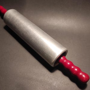 Può includere: Un mattarello d'argento con manici in legno rosso. Il mattarello è in metallo e ha una superficie liscia. I manici sono in legno e hanno una superficie testurizzata.