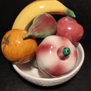 Peut inclure: Un bol de fruits en céramique avec une banane jaune, une poire à bout vert, une pomme rouge, une orange et une pêche aux accents rouges. Les fruits sont disposés dans un bol blanc texturé.