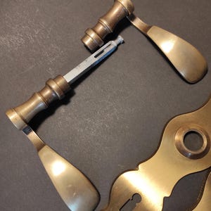 May include: An assortment of antique brass door hardware, including two door handles, a connecting rod, and a decorative escutcheon plate. The handles have a curved design and a tarnished finish, suggesting age and use. The hardware is arranged on a dark surface.