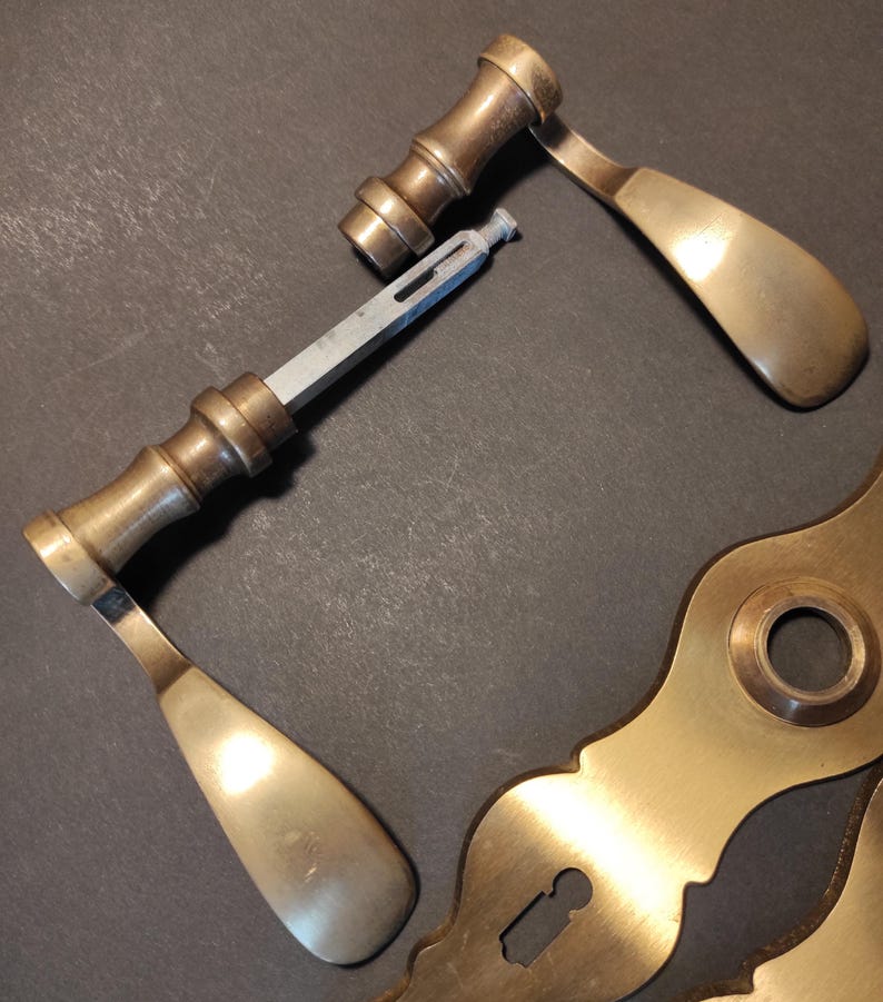 May include: An assortment of antique brass door hardware, including two door handles, a connecting rod, and a decorative escutcheon plate. The handles have a curved design and a tarnished finish, suggesting age and use. The hardware is arranged on a dark surface.
