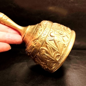 May include: A gold-colored metal hand bell featuring an ornate embossed pattern of leaves and birds. The bell has a long, slender handle with decorative carvings and the text JOHANNES clearly visible on the upper section of the bell body.