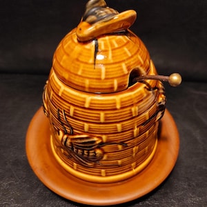 May include: A ceramic honey pot with a lid shaped like a hat, and a small spoon. The pot is brown with a beehive design and a bee relief. The pot sits on a matching saucer.