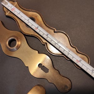 May include: Close-up of antique brass door hardware, including a keyhole plate and a decorative backplate. A measuring tape shows the backplate is approximately 8 inches long. The hardware has a warm, aged patina.