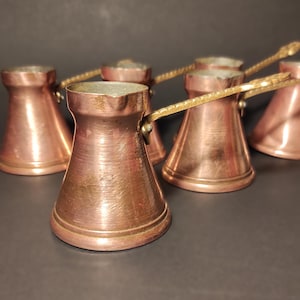 以下が含まれることがあります： 真鍮の取っ手が付いた銅製のコーヒーポットのコレクション。ポットはクラシックな先細りの形状で、磨かれた仕上げです。取っ手は装飾的で、デザインにエレガントさを加えています。ポットは暗い表面に配置されています。