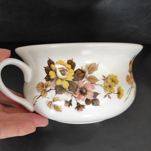 May include: White ceramic chamber pot with a floral design featuring yellow, pink, and brown flowers. The pot has a handle on the side.