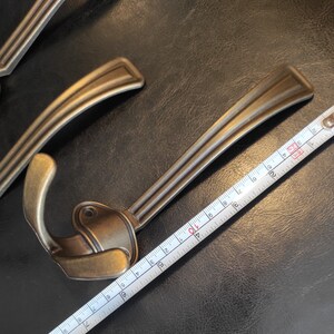 以下が含まれることがあります： 装飾的なデザインが施されたアンティークな真鍮製のフックが2つ。フックはメジャーで測定されています。