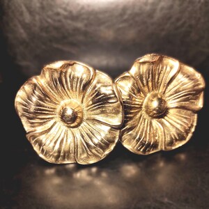 以下が含まれることがあります： 5枚の丸みを帯びた花びらを持つ、ゴールドトーンの金属製フラワーモチーフのペア。各花びらには放射状の細かな線が刻まれ、中央には滑らかで盛り上がった円形の中心部があります。金属の表面が光を反射し、花のデザインを際立たせています。