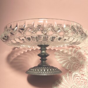 May include: A clear glass pedestal dish featuring a scalloped rim and a textured, pressed pattern. The dish is supported by a dark, tarnished metal base with a decorative, beaded edge design, providing a stable foundation for the glass bowl.