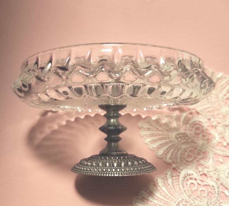 May include: A clear glass pedestal dish featuring a scalloped rim and a textured, pressed pattern. The dish is supported by a dark, tarnished metal base with a decorative, beaded edge design, providing a stable foundation for the glass bowl.