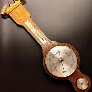 Puede incluir: Estación meteorológica de madera con termómetro, barómetro e higrómetro. La esfera del barómetro indica "Schön" y "Veranderliche". El termómetro está en grados Celsius. La madera tiene un tono marrón cálido.