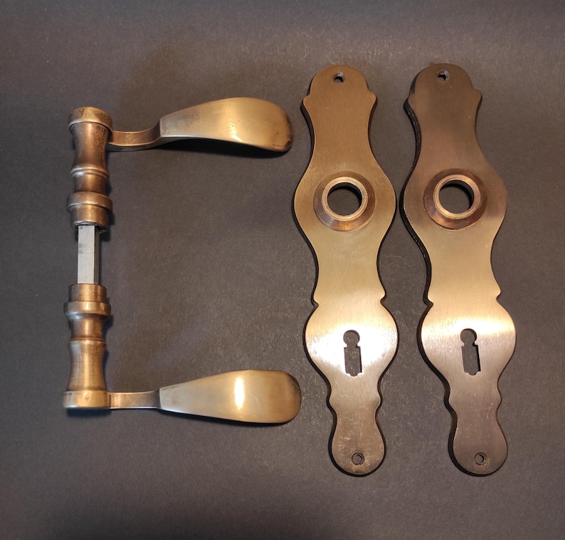 May include: Antique brass door hardware set, including a pair of door handles and two matching escutcheons. The handles have a curved design, and the escutcheons feature keyhole openings. The hardware has a vintage aesthetic.