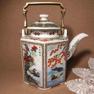 Op de afbeelding: Een decoratieve porseleinen theepot met een goudkleurige handgreep. De zeshoekige theepot is versierd met kleurrijke illustraties van vogels, bloemen en bomen. Het deksel heeft een groen en bloemenpatroon.