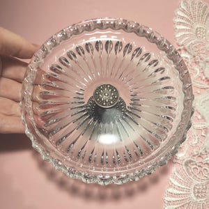 May include: A clear glass dish featuring a radial sunburst pattern of elongated indentations extending from the centre. The centre of the dish is adorned with a circular, metallic silver-toned knob that has a textured, dotted surface finish.