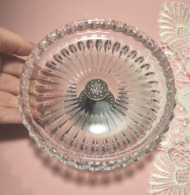 May include: A clear glass dish featuring a radial sunburst pattern of elongated indentations extending from the centre. The centre of the dish is adorned with a circular, metallic silver-toned knob that has a textured, dotted surface finish.