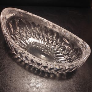 May include: A clear, oval-shaped crystal bowl with a detailed leaf pattern. The bowl is made of thick glass and has a slightly curved rim. The bowl is empty and sits on a dark surface.