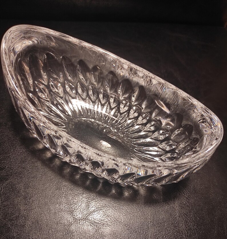 May include: A clear, oval-shaped crystal bowl with a detailed leaf pattern. The bowl is made of thick glass and has a slightly curved rim. The bowl is empty and sits on a dark surface.