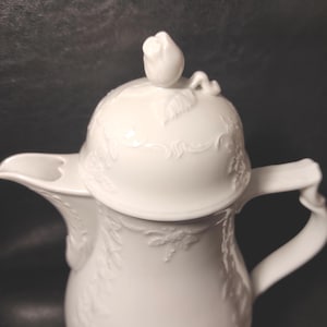 May include: A white porcelain jug with a lid, adorned with a rose bud and embossed floral designs. The jug has a curved spout and a handle, suitable for serving drinks. The design is elegant and classic, suitable for any occasion.