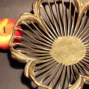 Könnte beinhalten: Eine goldfarbene Metallschale mit einem gewellten, durchbrochenen Design, das aus dünnen, strahlenförmig angeordneten vertikalen Lamellen besteht und eine blumenartige Form bildet. Ein roter und gelber Apfel liegt daneben.