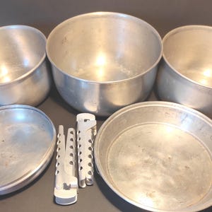 May include: A set of silver-colored aluminum camping cookware consisting of three round pots of varying sizes, one round lid, one round pan, and two detachable metal pot handles with perforated designs for gripping the cookware edges.