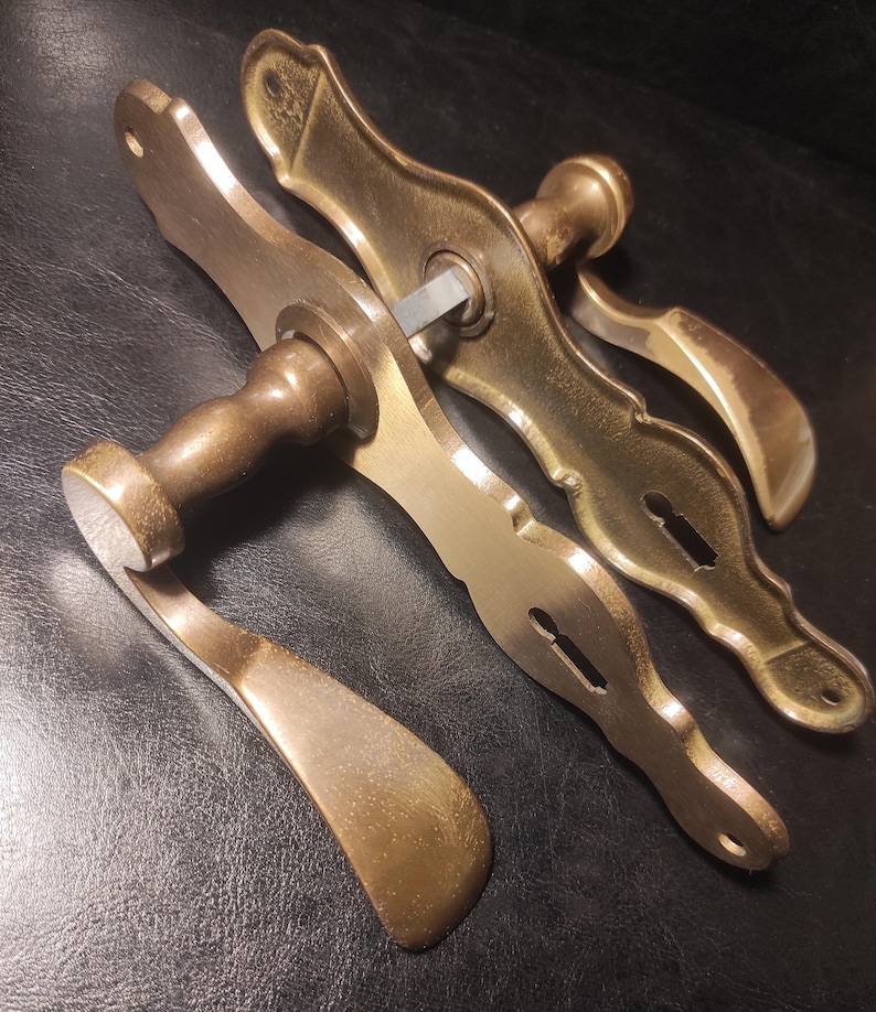 May include: Antique brass door handles with a curved design. The handles have a keyhole and a lever-style handle. The brass has a warm, aged patina. The door handles are likely for a vintage or antique door.