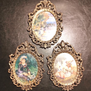 Puede incluir: Tres placas de pared ovaladas con marcos dorados metálicos ornamentados con volutas intrincadas y detalles florales. Cada marco contiene una escena artística impresa diferente, que incluye un paisaje, una persona en un jardín y un grupo.