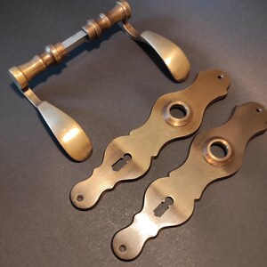 May include: Antique brass door handles and escutcheons. The handles have a curved design with a detailed grip. Two escutcheons with keyholes and circular openings are also included. The hardware has a warm, aged appearance.