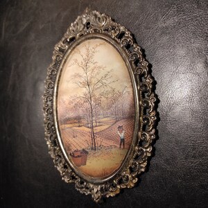 May include: An oval-shaped picture frame with an ornate silver finish. The frame contains a vintage painting of a rural landscape with a single figure in the distance.
