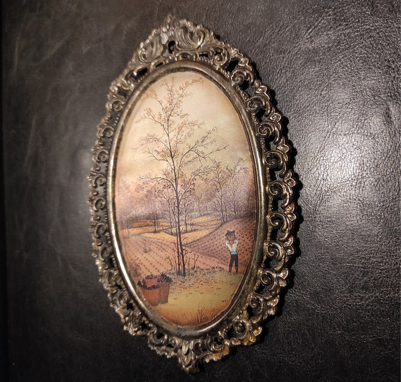 May include: An oval-shaped picture frame with an ornate silver finish. The frame contains a vintage painting of a rural landscape with a single figure in the distance.
