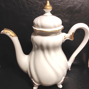 Op de afbeelding: Witte porseleinen theepot met gouden details op de tuit, het handvat en de knop. De theepot heeft een gegroefde body en staat op drie kleine voetjes. Het deksel is wit met gouden rand.
