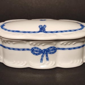 Könnte beinhalten: Weiße Porzellan-Schmuckschatulle mit einem blauen Seil- und Schleifendesign. Die Schachtel hat einen gewellten Rand und einen Deckel mit einem erhabenen Seildesign.