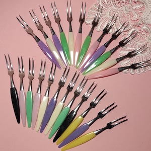 May include: A collection of vintage dessert forks with stainless steel tines and colorful plastic handles. The handles are in various pastel colors including pink, green, purple, yellow, and white. The forks are arranged on a pink surface.