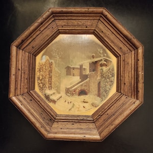 Puede incluir: Una pintura enmarcada que representa una escena de pueblo nevado con edificios de piedra y algunas aves en primer plano. El marco es octogonal y de madera oscura.