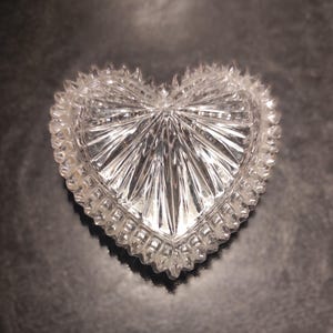 May include: A clear, heart-shaped trinket box with a textured, faceted design. The box has a scalloped edge and a starburst pattern radiating from the center. The box is made of glass or crystal.