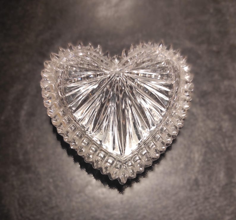 May include: A clear, heart-shaped trinket box with a textured, faceted design. The box has a scalloped edge and a starburst pattern radiating from the center. The box is made of glass or crystal.