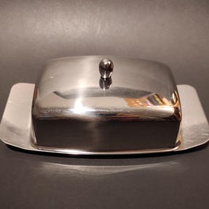 Puede incluir: Un mantequillero de plata con tapa y un pomo redondo en la parte superior. El mantequillero está hecho de acero inoxidable y tiene un acabado brillante.