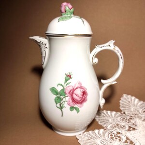 Puede incluir: Cafetera de porcelana blanca con un diseño de rosa rosa. La cafetera presenta un asa curva, un pico y una tapa rematada con un capullo de rosa rosa. Los adornos dorados acentúan el borde y el asa.