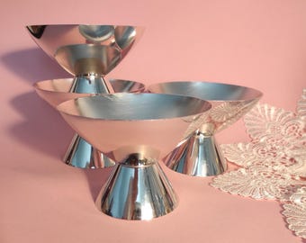 May include: A set of three silver-colored metal dessert cups with wide, shallow bowls and conical pedestal bases. The polished surfaces reflect light, creating a bright, metallic appearance. The cups are arranged in a cluster on a flat surface.