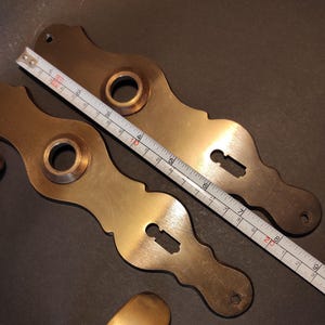 May include: Two antique brass door handle backplates with keyhole cutouts and a circular opening. A ruler shows the backplates are approximately 8 inches long. The brass has a warm, aged patina.