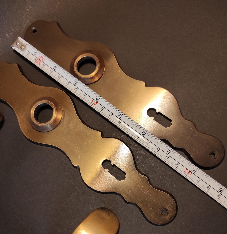 May include: Two antique brass door handle backplates with keyhole cutouts and a circular opening. A ruler shows the backplates are approximately 8 inches long. The brass has a warm, aged patina.