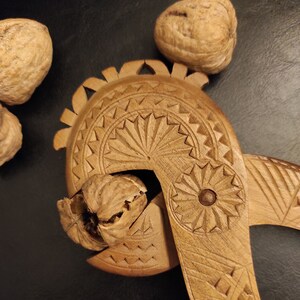 Op de afbeelding: Een houten notenkraker met ingewikkelde houtsnijwerk. De notenkraker is open en er zit een walnoot in. Er liggen drie walnoten op de tafel.