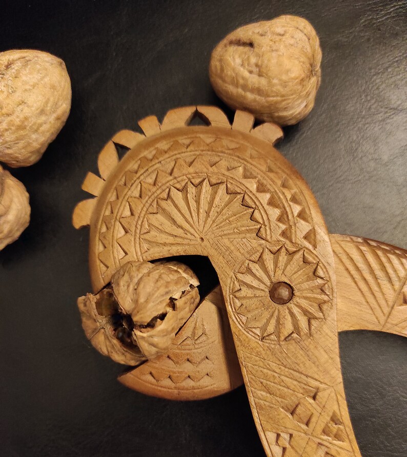 Op de afbeelding: Een houten notenkraker met ingewikkelde houtsnijwerk. De notenkraker is open en er zit een walnoot in. Er liggen drie walnoten op de tafel.