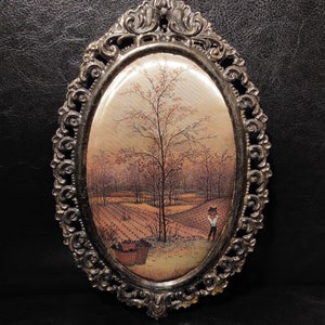 May include: An oval-shaped framed print of a painting depicting a rural landscape with a lone figure in the foreground. The frame is ornate and silver in color.
