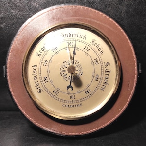May include: A vintage barometer with a brown leather frame and a cream-colored face. The face features a needle pointing to the current atmospheric pressure, with text in German. The word "GOLDTIME" is printed at the bottom.